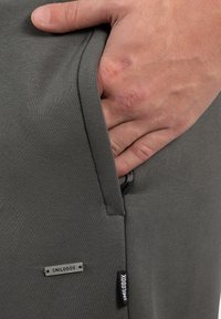 Grå stof joggers har en hånd i en sidelomme, et lille metalmærke med prægningen "SMILODOX" og en lynlåslomme-detalje.