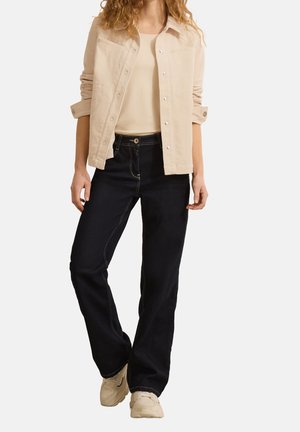 Mujer que lleva una chaqueta beige con botones sobre un top beige, vaqueros negros de pierna ancha con costuras en contraste y zapatillas beige, de pie con una pierna hacia adelante.