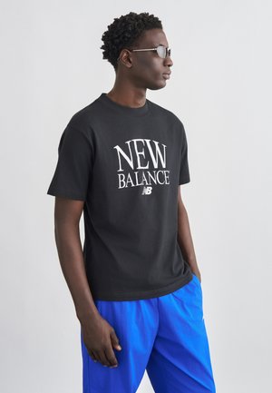 Muž v čiernom tričku New Balance, modrých nohaviciach a slnečných okuliaroch stojaci bokom s jednou rukou v vrecku na jednofarebnom pozadí.