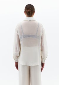 Lekka, przejrzysta biała koszula z kołnierzykiem i bufiastymi rękawami, z subtelnie widoczną niebieską warstwą pod spód oraz teksturowanym, falującym materiałem.