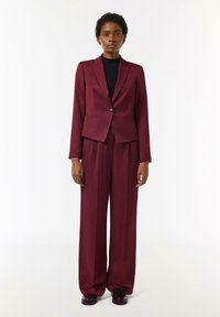 Burgundowy garnitur z dopasowaną marynarką na jeden guzik oraz spodniami o szerokich nogawkach. Materiał wydaje się gładki z subtelną teksturą.