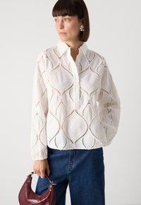 Blouse blanche à manches longues avec un motif en dentelle délicat et une patte de boutonnage. Assortie à un jean bleu et un petit sac à main bordeaux.
