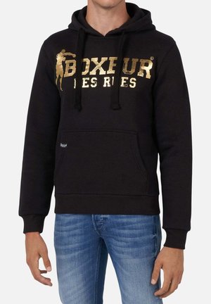 Uomo con felpa nera con cappuccio con scritta dorata "Boxeur Des Rues" e figura da pugilato, abbinata a jeans blu, in piedi contro uno sfondo bianco.