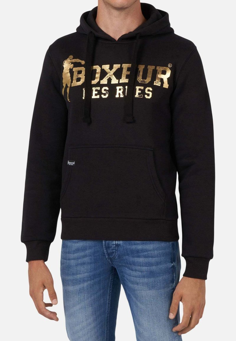 Homme portant un sweat à capuche noir avec un texte doré « Boxeur Des Rues » et une silhouette de boxeur, accompagné d'un jean bleu, debout devant un fond blanc.