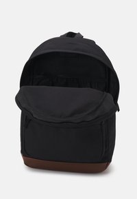 Sac à dos noir avec compartiment principal ouvert, poche avant et base marron, debout sur fond blanc.