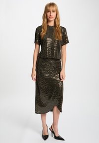 Femme portant un haut à manches courtes en sequins noirs et une jupe midi assortie avec des talons hauts pointus noirs, se tenant devant un fond blanc.