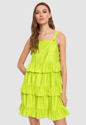 Donna che indossa un vestito a balze senza maniche di colore verde lime, in piedi davanti a uno sfondo grigio chiaro uniforme.