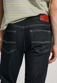 Tummansiniset farkut, joissa on kaksi takataskua, keltaista tikkausta ja ruskea nahkaleima, jossa näkyy brändin nimi. Slim fit -malleja.