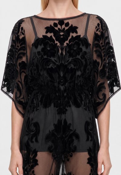 Woman wearing a black sheer dress with intricate velvet floral patterns and loose short sleeves, visible black undergarments underneath.