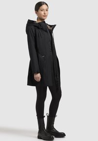 Schwarze wasserfeste Jacke mit Kapuze, ausgestattet mit Druckknöpfen, Seitentaschen und einem strukturierten Innenfutter. Kombiniert mit schwarzen Leggings und Stiefeletten.