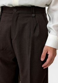 Bruine op maat gemaakte broek van een textuurstof, met twee voortuigen, plooien en een vlakke voorkant. Gecombineerd met een wit shirt.