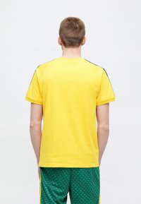 Żółta bawełniana koszulka z krótkimi rękawami, z zielonymi akcentami na ramionach, zaokrąglonym dekoltem i luźnym krojem.