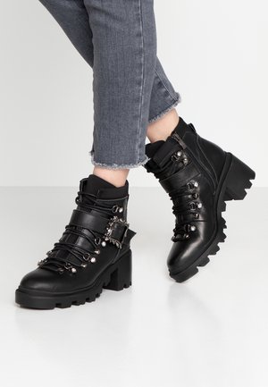 Bottines noires en cuir avec un talon épais, présentant des boucles décoratives, des accents métalliques et un design à lacets avec des semelles texturées.