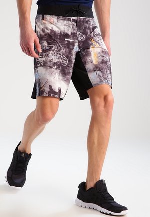 Mann trägt schwarz-weiß gemusterte Shorts mit abstraktem Muster und schwarze Sportschuhe, steht mit einem Bein leicht nach vorne gesetzt.