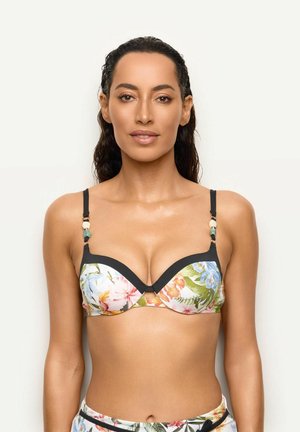 Kobieta z mokrymi włosami, ubrana w top bikini w kwiatowy wzór z czarną lamówką, stoi na jednolitym jasnym tle, zwrócona przodem.