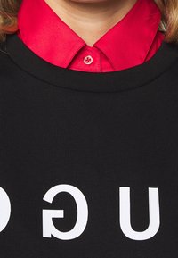 Felpa nera con grande testo bianco sul davanti. Presenta una camicia rossa con colletto visibile sotto, che mette in risalto il contrasto dei colori.