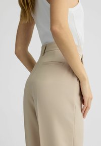 Pantaloni beige realizzati in tessuto morbido, design sartoriale e dettagli sulla tasca posteriore, abbinati a un top bianco aderente.