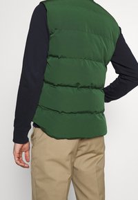 Vihreä toppaliivi, jossa on sileä pinta ja vaakasuora ompelu, kerrospukeutuneena tumman pitkähihainen paidan päälle, yhdistetty vaaleisiin housuihin.