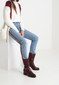 Brązowe skórzane kozaki za kostkę na słupkowym obcasie, ozdobione dekoracyjnymi ćwiekami z boku. W połączeniu z jasnoniebieskimi jeansami i białą koszulą.