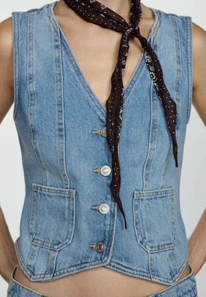 Mouwloze blauwe denimvest met voorzakken en metalen knopen, gedragen met een zwarte sjaal met patroon, geknoopt in een knoop.