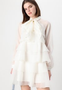 Rochie albă, stratificată, realizată dintr-un material ușor și transparent. Prezintă un guler înalt cu o fundă mare și mâneci lungi bufante.