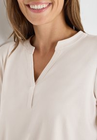 Top beige chiaro con colletto arrotondato e patta parziale. Tessuto dalla texture liscia con dettaglio di cucitura sottile lungo il collo.