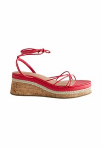 Sandale rouge en forme de coin avec plateforme en liège, fines lanières sur les orteils et lien à nouer autour de la cheville sur fond blanc.