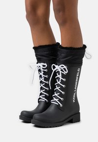 Black rubber boots with white laces, a soft black fabric cuff, and embossed "KARL LAGERFELD" branding on the side. Flat soles, knee-high design.