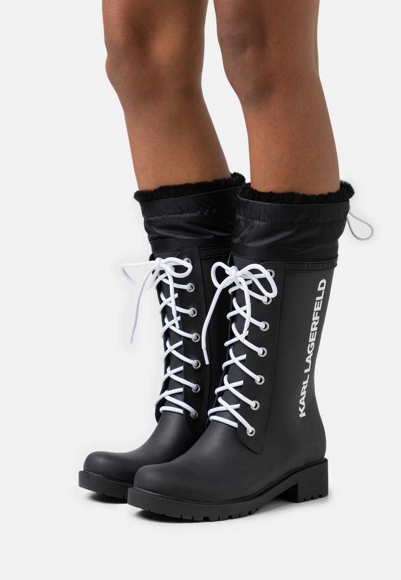 Black rubber boots with white laces, a soft black fabric cuff, and embossed "KARL LAGERFELD" branding on the side. Flat soles, knee-high design.