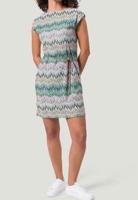 Kobieta w sukience w wielokolorowy wzór w zygzaki, z przewiązanym paskiem w talii i białych trampkach, stojąca z rękami za plecami.