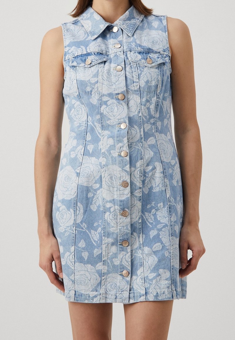 Robe en denim bleu clair sans manches avec motif floral blanc, boutonnée devant, col et deux poches à rabat sur la poitrine portée par une personne debout.