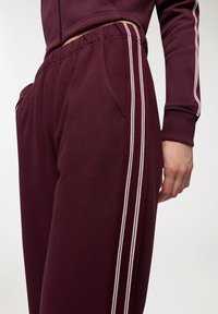 Burgundowe dresy o luźnym kroju, z białymi podwójnymi paskami po bokach, elastycznym pasem i kieszeniami bocznymi.