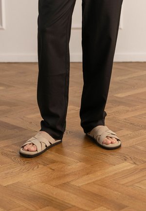 Persona con pantaloni neri e sandali beige intrecciati in piedi su pavimento in parquet di legno marrone chiaro.