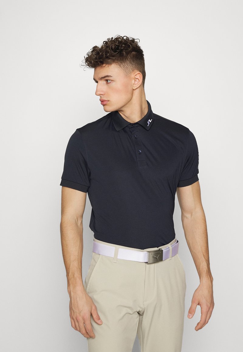 Camisa polo azul marino con diseño de cuello y tres botones, combinada con pantalones beige y un cinturón lila claro con hebilla de plata.