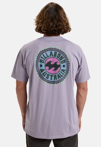 Camiseta de algodón morada con un gráfico circular en la parte trasera que dice "Billabong Australia", formas onduladas y acentos multicolores.