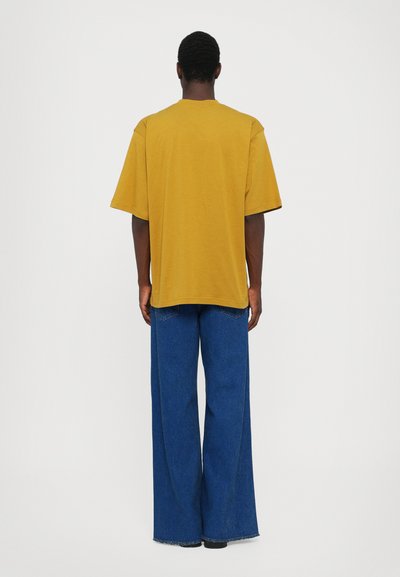 Un tricou galben supradimensionat cu mâneci scurte, purtat împreună cu blugi cu fason larg, de culoare albastră. Țesătura pare moale și are o croială lejeră.