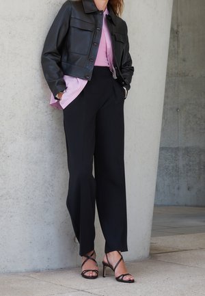 Vrouw die een zwarte cropped leren jas draagt, een roze blouse met knopen, zwarte wijde broek met hoge taille en zwarte sandalen met hoge hak en bandjes.