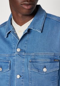 Dark-skinned person wearing light blue denim jacket with metal buttons and two chest pockets over a white buttoned top.
