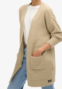 Beige ribbelgebreide cardigan met open voorkant, lange mouwen, twee voorkwaarten en een klein logo-opdruk in de onderste linkerhoek.