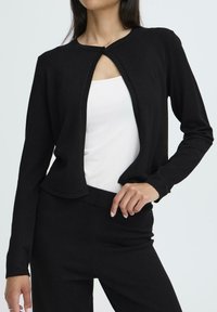 Schwarze Strickjacke mit Rundhalsausschnitt, offenem Vorderteil und einem einzigen Knopfverschluss, enganliegenden Ärmeln und einer weichen Textur, kombiniert mit einem weißen Oberteil.