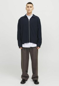 Marineblaue, gerippte Zip-Jacke mit großem Kragen, getragen über einem weißen Hemd. Lockere graue Hose und schwarze Schuhe vervollständigen das Outfit.