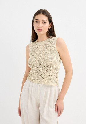 Top sin mangas beige con un patrón de diamantes, hecho de un material de punto texturizado y ligero; combinado con pantalones de color claro y de corte holgado.