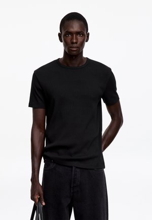 Homme à la peau foncée portant un t-shirt noir uni à manches courtes et un pantalon noir, debout avec une expression neutre sur fond blanc.