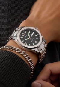 Montre en acier inoxydable argenté avec un cadran noir, des index horaires blancs, un affichage de date et un bracelet métallique poli.