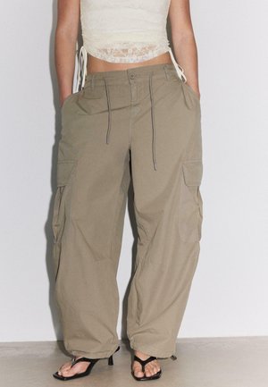 Femme portant un pantalon cargo ample kaki avec des poches, un crop top en dentelle blanche et des sandales noires à talons, debout devant un fond uni.
