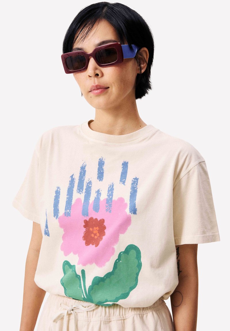 Persona con gafas de sol rectangulares moradas y azules, y una camisa beige con un diseño grande de flor rosa y pinceladas azules.