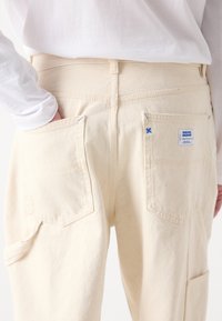 Pantalon cargo beige à coupe droite, doté de deux poches arrière et d'accents de coutures bleues. La texture du tissu est légèrement brossée.
