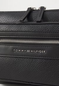 Černá texturovaná kožená taška s dvěma stříbrnými zipy, s gravírovaným logem "TOMMY HILFIGER" a elegantními designovými detaily.