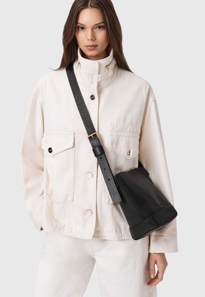 Sac bandoulière en cuir noir avec une forme structurée, présentant une finition texturée et une sangle ajustable, associé à une veste crème.