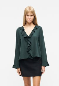 Ξανθιά γυναίκα φορώντας μια σκούρα πράσινη μπλούζα με βολάν και μακριά μανίκια, καθώς και μια μαύρη μίνι φούστα, στέκεται μπροστά σε ένα απλό λευκό φόντο.